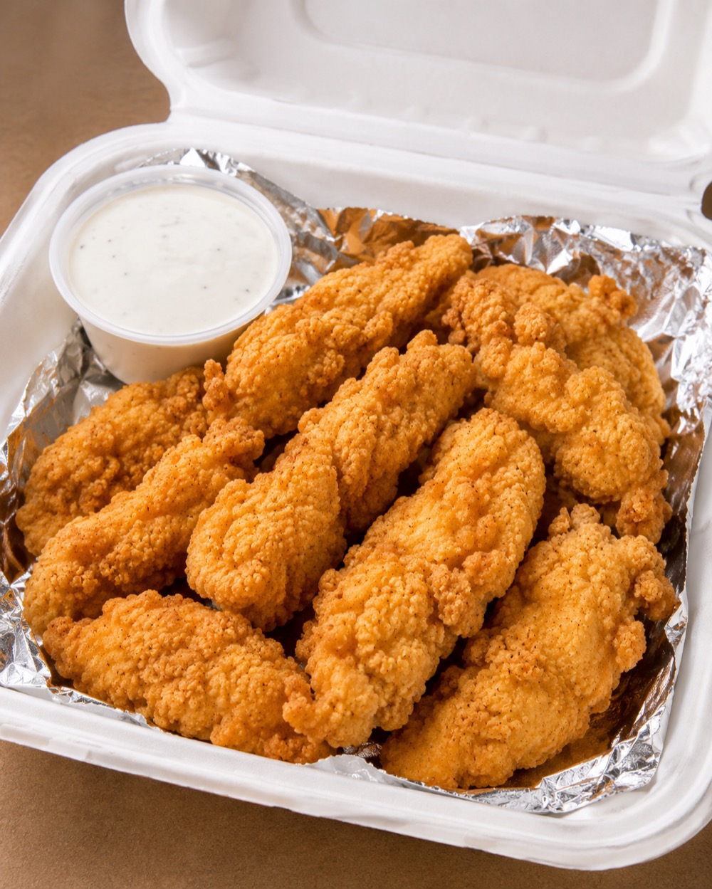 Hand-battered golden-fried chick'n tenders in a foil-lined takeout box with a cup of ranch.