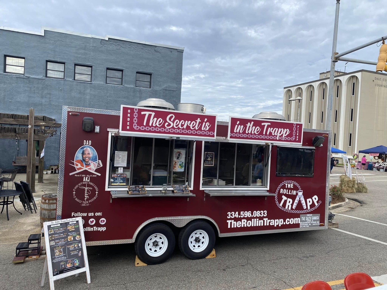 The Rollin' Trapp trailer parked in downtown Ozark, its burgundy wrap reading The Secret is in the Trapp, with the memorial portrait of the owners' father and a chalkboard menu beside the service window.